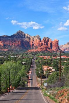 an empty road in the desert with red rocks behind it and green trees on both sides