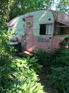 an old pink trailer is parked in the middle of some bushes and trees, with a small table next to it