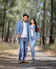 a man and woman standing in the middle of a dirt road with trees behind them