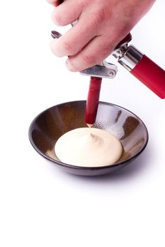 a person is pouring cream into a bowl with a red spatula in it on a white background