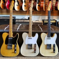 three guitars are lined up next to each other in front of guitar cases and amps