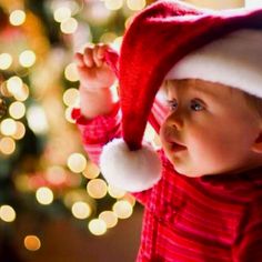 a baby wearing a santa hat next to a christmas tree with lights in the background