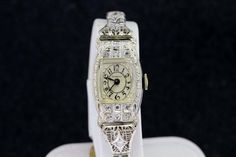 This wrist watch is a Robbins's that is 14K gold one piece case that has pave diamonds set around the watch.The diamonds are estimated to be a SI2 clarity and is a G/H color.The band is hinged and has filigree and pave set diamonds set on it. The face of the dial is engraved with daisies and leaves and has a domed crystal. The case of the watch is engraved with flowers and various other floral patterns.It has a clasp and safety latch to make it secure on the wrist. The case of the watch measures Timeless Diamond Wedding Watches, Diamond Wedding Watch With Diamond Hour Markers, Evening Diamond Watch With Art Deco Style, Wedding Diamond Watch With Diamond Hour Markers, Evening Art Deco Diamond Watch With Accents, Diamond White Wedding Watch With Diamond Hour Markers, Timeless Diamond Watch For Wedding, Formal Diamond White Watch With 17 Jewels, Wedding Diamond Watch With Diamond Accents