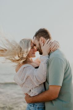 a man and woman hugging on the beach with their hair blowing in the wind as they look into each others eyes