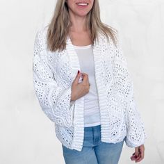 a woman wearing a white crochet cardigan and jeans standing in front of a white wall