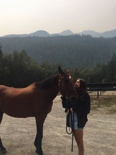 a woman standing next to a brown horse on top of a dirt field with mountains in the background