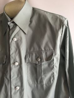 Awesome vintage men's uniform work shirt jacket. The military inspired cut and color encourage me to believe this was a aviation uniform of some sort. Somewhere between a shirt and a jacket, these piece has some great details. The material is most likely a permanent press style poly/cotton blend, though no content label is present. The shirt has a button front styling much like a classic men's dress shirt but with a more boxy cut and elastic waist. It has small loops and holes near top shoulder Retro Solid Color Shirt With Button Closure, Retro Solid Shirt With Button Closure, Military Style Button-up Work Shirt, Military Style Button-up Shirt For Work, Military Style Shirt For Work With Button Closure, Military Style Workwear Button-up Shirt, Long Sleeve Workwear Uniforms, Military Long Sleeve Work Shirt, Military Style Shirt With Buttons For Work