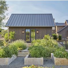 a small house with lots of plants and flowers in the front yard, surrounded by stone planters