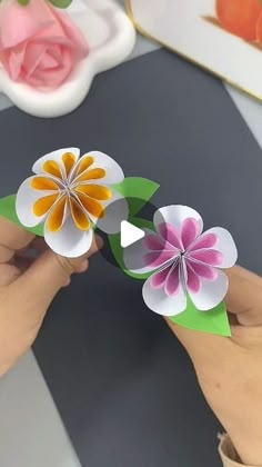 two paper flowers being held by someone's hand on top of a black and white table