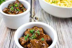 two white bowls filled with meat and pasta on top of a wooden table next to another bowl