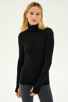 A person with long, light-colored hair is wearing a black Jackson Rib Full Length Turtleneck from SPLITS59 and gray bottoms, standing against a plain white background. Fitted Winter Tops With Thumbholes, Winter Fitted Tops With Thumbholes, High Stretch Black Winter Tops, High Stretch Tops With Ribbed Cuffs For Fall, Black Tops With Ribbed Cuffs, Versatile Top With Ribbed Cuffs And Stretch, Versatile Stretch Top With Ribbed Cuffs, Versatile Tops With Ribbed Cuffs And Stretch, Stretch High Neck Tops With Ribbed Cuffs