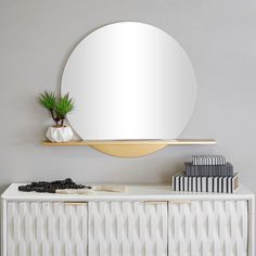 a white and gold dresser with a round mirror on the wall above it, next to a plant