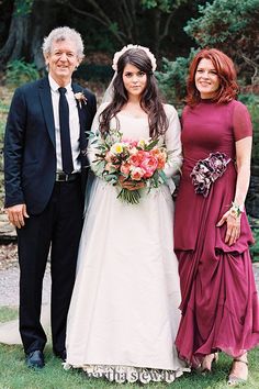 two women and a man standing next to each other in front of some trees with flowers