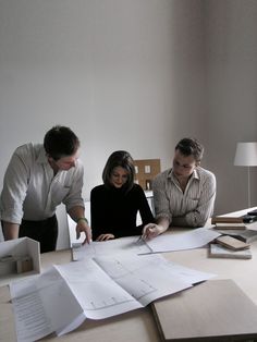 three people sitting at a table with papers on it and one person standing over them