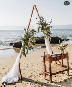 a wooden table topped with flowers next to an arch on top of a sandy beach