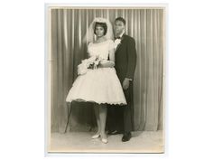 an old black and white photo of a man and woman dressed in wedding attire standing next to each other