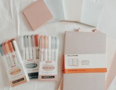 some pens and notebooks are laying out on a bed with white sheets, pink paper, and orange ribbon