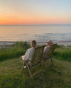 two women sitting on lawn chairs watching the sunset at the beach with their backs to each other