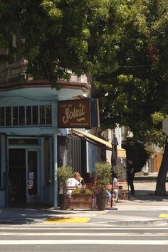 people are sitting at tables on the sidewalk in front of a small store with an awning