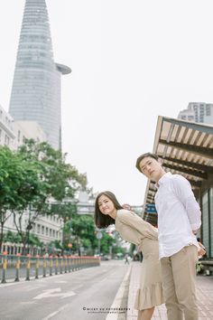 a man and woman standing on the sidewalk in front of a tall building with a sky scraper behind them