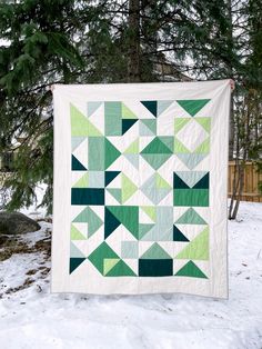 a green and white quilt hanging from the side of a tree in the snow with trees behind it