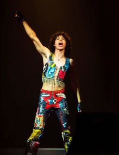 a man with his arms in the air while standing on top of a stage wearing colorful pants