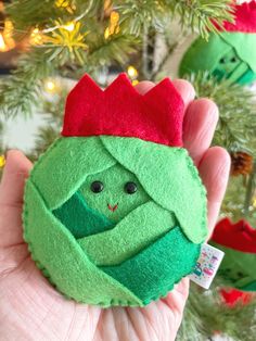 a hand holding a green toy with a red crown on it's head in front of a christmas tree