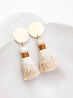 two white tasseled earrings with gold disc and brown ribbon on top, sitting on a plate