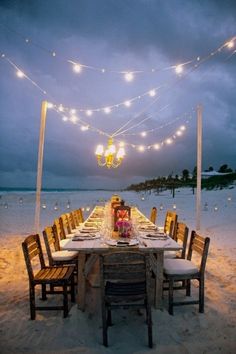 an outdoor dining table set up on the beach at night with lights strung over it