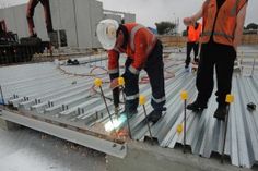 two men in orange vests working on a metal structure with other workers standing around