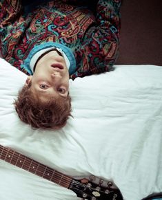 a young man laying on top of a bed next to an electric guitar and looking up at the camera