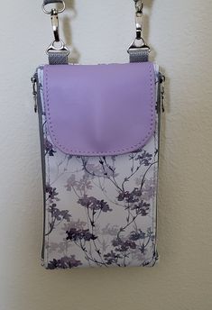 The vinyl exterior fabric on this unique bag features delicate lavender and gray flowers. This modified Zip n Go can be a crossbody or a shoulder bag or a wristlet or a fanny pack/waist bag or a wallet. It has zippered compartments on both sides. One has card slots, a paper money area, and a zippered compartment for change.  The other will hold a cell phone or miscellaneous "stuff." In addition, the front magnetic snapped flap conceals another pocket that is great for a cell phone. The long strap is gray webbing and is adjustable and removable. It also comes with a wristlet strap. Very unique, functional bag with a lavender front flap, silver hardware, and silver water-resistant lining. Purple Crossbody Phone Bag With Cell Phone Pocket, Purple Travel Phone Bag With Removable Pouch, Purple Phone Bag With Removable Pouch, Purple Phone Bag With Removable Pouch For Daily Use, Rectangular Purple Phone Bag With Removable Pouch, Vinyl Exterior, Silver Water, Wallet Wristlet, Convertible Bags