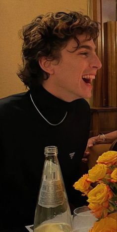 a young man sitting at a table with flowers in front of him and a bottle of wine