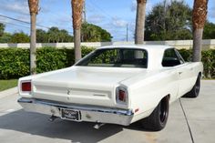 an old white car parked in a driveway next to some palm trees and bushes on a sunny day