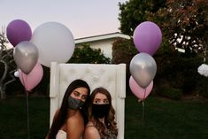 two women sitting on a chair with balloons in the air and one wearing a face mask