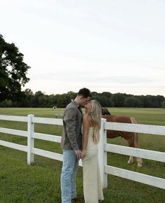 a man and woman kissing in front of a white fence with a horse behind them