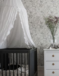 a baby crib next to a white dresser and wallpaper with flowers in vases