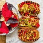 three hot dogs with toppings next to a bowl of chips