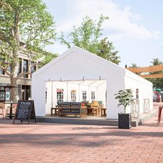 a white tent with tables and chairs in front of it on a brick patio next to trees