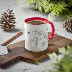 a red and white coffee mug sitting on top of a wooden cutting board next to pine cones