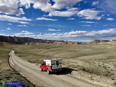 a red truck driving down a dirt road in the middle of desert with blue sky and clouds