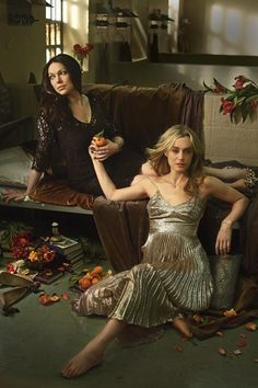 two women sitting on a couch in front of a table full of flowers and fruit