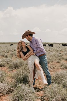 a man and woman standing in the middle of an open field with cows behind them