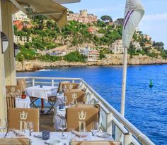 an outdoor dining area with tables and chairs on the deck overlooking the water in front of some buildings