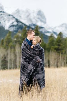 a man and woman wrapped in a blanket standing in tall grass with mountains in the background