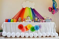 a table topped with lots of white balloons and rainbow colored streamers next to a cake