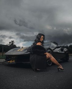 a woman sitting on the ground next to a black car with clouds in the background