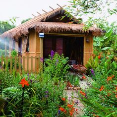 a small house with a thatched roof surrounded by plants and flowers in the foreground