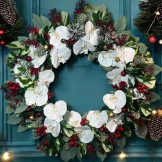 a christmas wreath with white flowers and greenery hanging on a blue door surrounded by pine cones