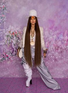 a woman with long brown hair standing in front of a purple wall wearing a white hat